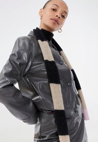 Veste en cuir grise avec une finition brillante, dotée de gros boutons et de deux poches, accompagnée d'une écharpe en tricot rayée noire, beige et rose.