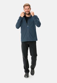 Heren blauwe softshell jas met een rits aan de voorkant, verstelbare kraag en twee zijzakken, gecombineerd met zwarte broeken en grijze schoenen.