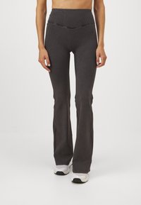 Kvinna som bär högmidjade, ribbade, mörkgrå flare leggings tillsammans med vita sneakers, stående framför en enkel vit bakgrund.