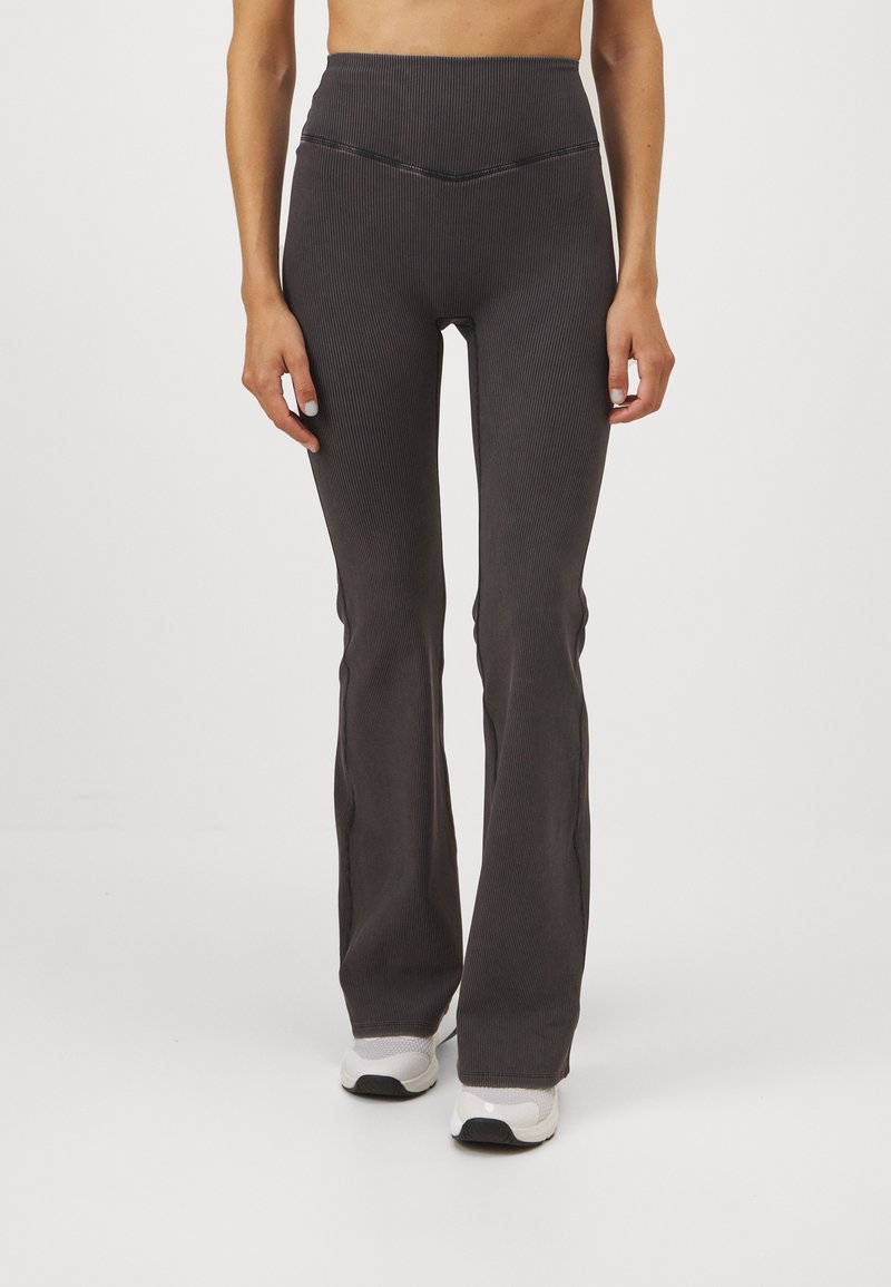 Kvinna som bär högmidjade, ribbade, mörkgrå flare leggings tillsammans med vita sneakers, stående framför en enkel vit bakgrund.