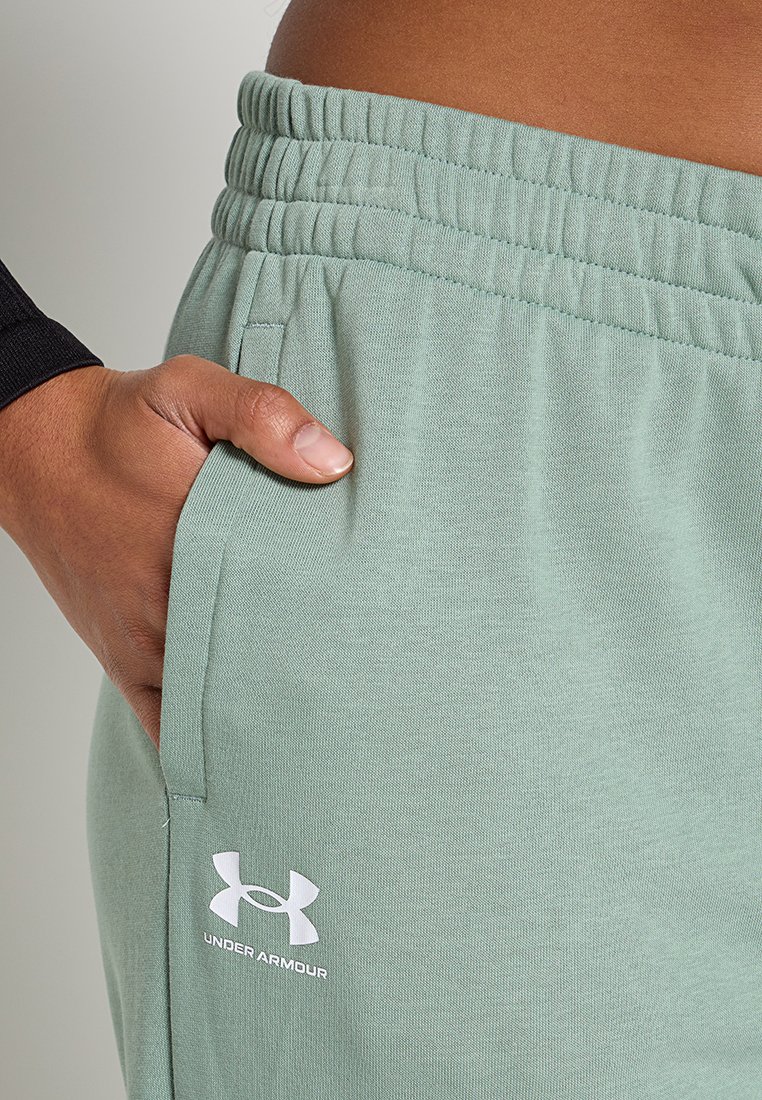 Ręka w kieszeni jasnooliwkowych spodni dresowych Under Armour z elastycznym paskiem, na osobie ubranej w czarny długi rękaw.