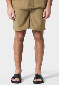 Shorts beiges à finition lisse, design jusqu'aux genoux, et sandales noires à enfiler avec une tige texturée. Accent sur le confort et la praticité.