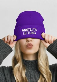 Purple knitted beanie with a rectangular patch displaying "ANSTALTS LEITUNG" in white text, worn low over the forehead. Textured fabric detail.