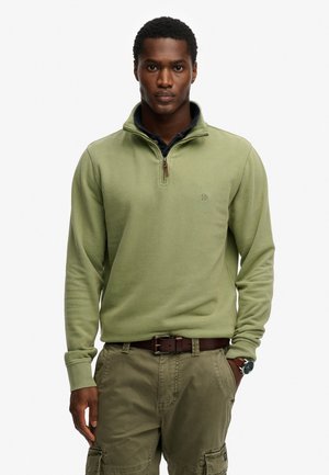 Mężczyzna w jasnozielonym pulowerze z krótkim zamkiem na tacę, noszący ciemną koszulę, khaki spodnie typu cargo i brązowy pasek, stojący z jedną ręką w kieszeni.