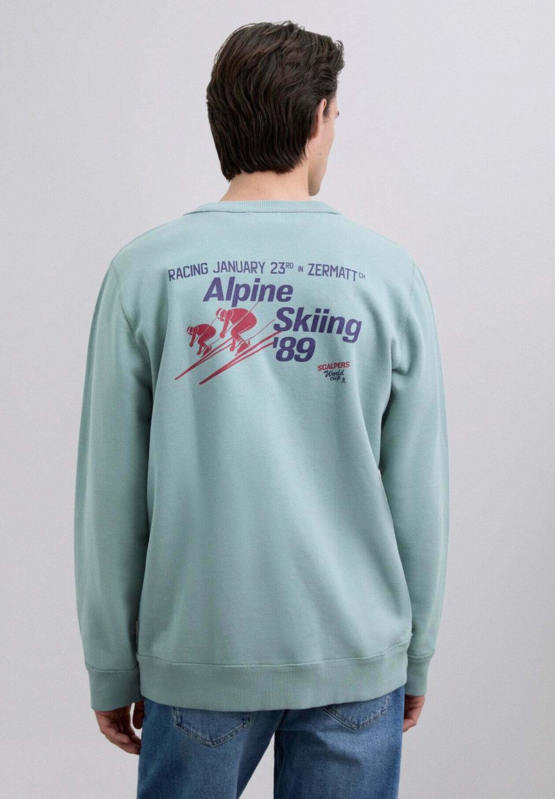 Sudadera verde menta con cuello redondo, que presenta una impresión gráfica en la parte posterior con figuras de esquí y texto en púrpura y rojo.