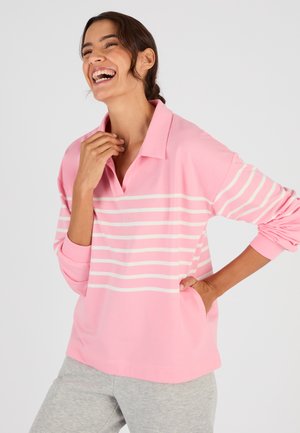 Donna sorridente che indossa una camicia a righe rosa e bianca a maniche lunghe con collo e pantaloni grigio chiaro, in piedi contro uno sfondo semplice.