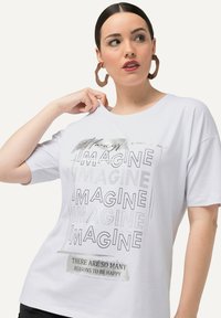 Hvid kortærmet T-shirt med et grafisk print, der viser ordet "IMAGINE" i forskellige stilarter samt et slogan: "DER ER SÅ MANGE GRUNDE TIL AT VÆRE GLAD."