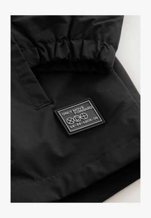 Melns jaka ar detaļām, kurās ir gumijas uzlīme ar tekstu "VIEGLI PĀRVIESIES TIKAI UZ PRIEKŠU" un ģeometriskām akcentiem, izgatavota no mīksta teksturēta auduma.