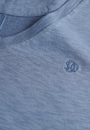 T-shirt bleu clair à manches courtes avec un col en V. Il présente un tissu texturé et un logo brodé dans le coin supérieur droit.