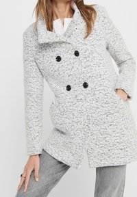 Cappotto doppiopetto grigio chiaro con superficie texturizzata, grandi bottoni neri e tasche laterali. Presenta un colletto alto e maniche lunghe.