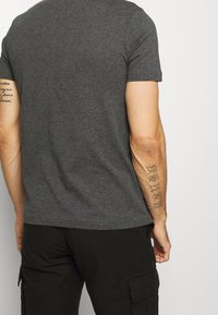 Hombre con una camiseta gris oscuro y pantalones cargo negros, mostrando tatuajes en ambos antebrazos, de pie contra un fondo claro y liso.