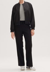 Czarna kurtka typu bomber z elastycznymi mankietami, na czarno-białym paskowanym topie, w zestawieniu z czarnymi spodniami i białymi trampkami. Widoczne szczegóły tekstury materiału.