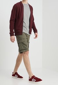 Cardigan bordeaux con zip, t-shirt grigia, pantaloni cargo verde oliva e sneakers in suede rosse con strisce bianche. Modello in piedi contro uno sfondo chiaro.