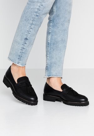 Mocasines de cuero negro con parte superior texturizada, detalle de borlas y suela resistente. Combinados con jeans de mezclilla desgastados de color azul claro para crear contraste.