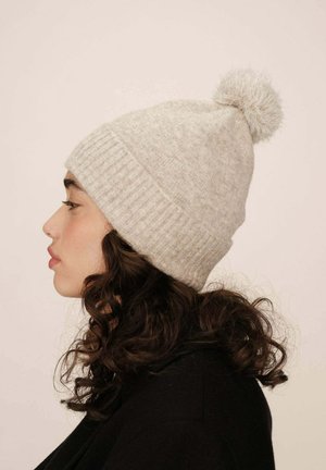 Bonnet tricoté gris clair avec un design côtelé et un pompon duveteux sur le dessus, porté par une personne aux cheveux bouclés, vue de profil.