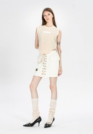 Mujer con el pelo largo y marrón, que lleva una parte superior sin mangas beige, falda blanca con cordones y bolsillos, calentadores de piernas beige arrugados y tacones puntiagudos negros.