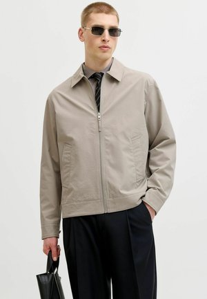 Ung mand iført beige jakke med lynlås, sorte bukser, solbriller, holder en sort lædertaske, står mod en ensfarvet lys baggrund.