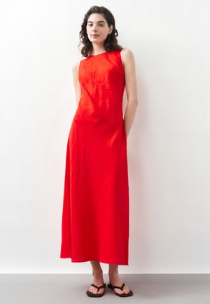 Donna in piedi contro un muro bianco che indossa un vestito lungo rosso acceso senza maniche e infradito nere, con le mani dietro la schiena.