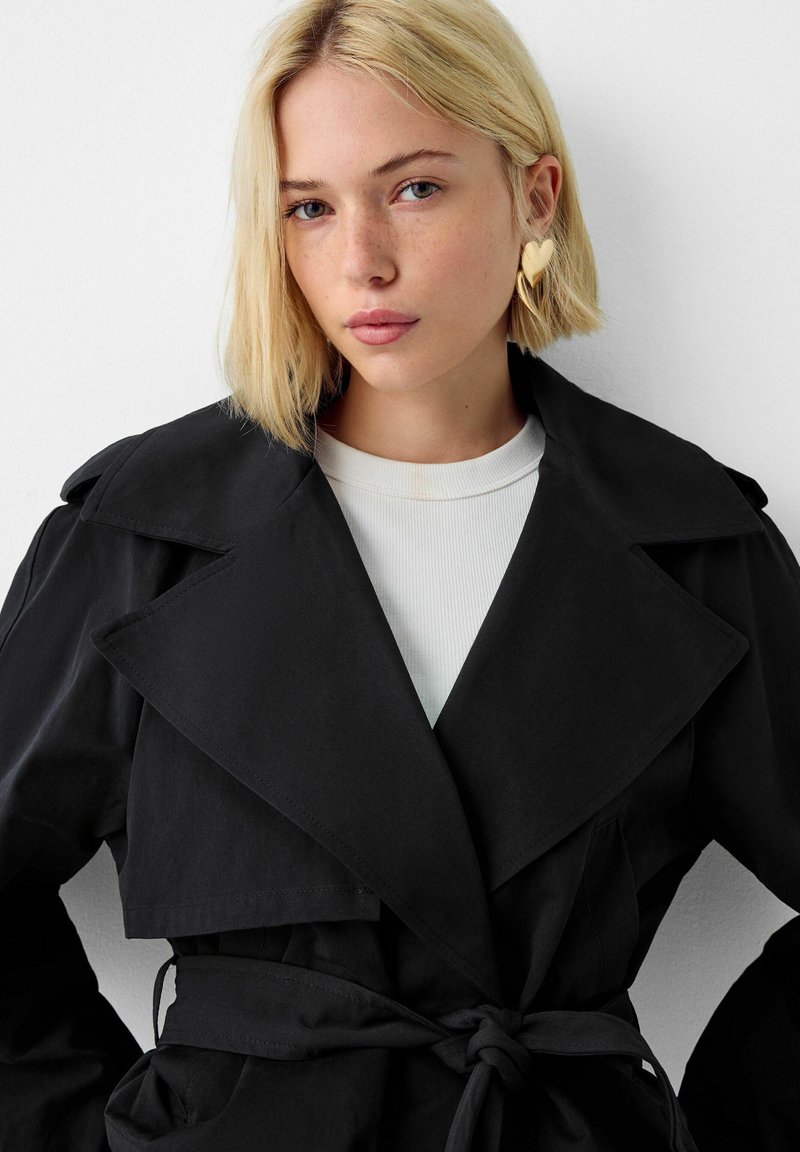 Cappotto nero con colletto a v, vita cinturata e tasche larghe. Il tessuto è liscio con una leggera lucentezza.