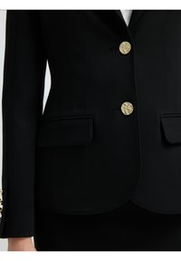 Blazer negro con botones dorados, dos bolsillos frontales, silueta entallada y detalles de costura sutiles. Tejido suave con un diseño estructurado.