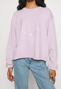 Kvinna iklädd en ljuslila sweatshirt med en glänsande cirkulär virveldesign, kombinerad med ljusblå jeans.