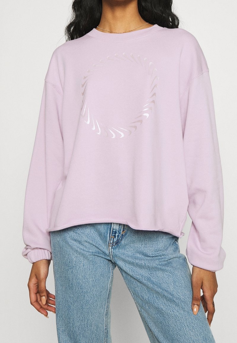 Kvinna iklädd en ljuslila sweatshirt med en glänsande cirkulär virveldesign, kombinerad med ljusblå jeans.