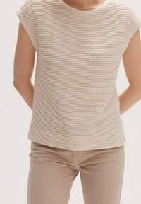 Cremfarbener, strukturierter Oberteil mit horizontalen Wellen und kurzen Ärmeln, kombiniert mit beigen Hosen. Einfaches Design, weicher Stoff, keine sichtbaren Hardware.