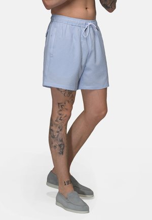 Man draagt lichtblauwe seersucker korte broek met trekkoord en grijze slip-on schoenen, toont tatoeages op zijn linkerbeen.