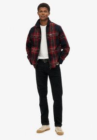 Chaqueta de franela a cuadros rojos y marinos con dos bolsillos en el pecho, llevada sobre una camiseta blanca. Combinada con pantalones negros y zapatillas beige.