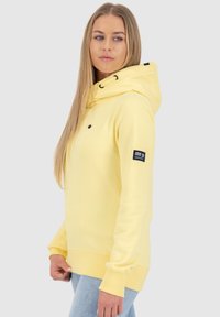 Jonge vrouw met lang blond haar, gekleed in een lichtgele hoodie met capuchon en lichtblauwe jeans, staande tegen een effen achtergrond.