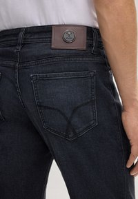 Jean en denim foncé avec une texture lisse, doté d'une patch en cuir marron avec un logo en relief et de doubles poches arrière avec des coutures discrètes.