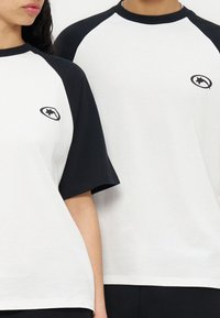 Δύο άτομα φορούν λευκές μπλούζες με μαύρα μανίκια raglan και ένα μικρό μαύρο λογότυπο αστέρι στο στήθος, στέκονται δίπλα δίπλα.