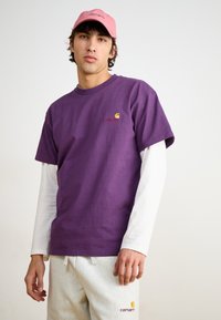 Camiseta de algodón morada con cuello redondo, superpuesta a una camisa de manga larga blanca; presenta el logo rojo de Carhartt en el pecho. Pantalones de chándal gris claro.