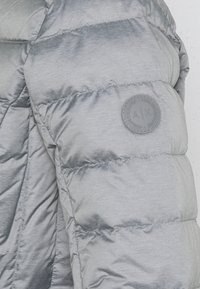 Harmaa puffatakki, jossa on virtaviivainen muotoilu, kiiltävä pinta, vaakasuora tikkikaulus ja pyöreä logopatchi hihassa.