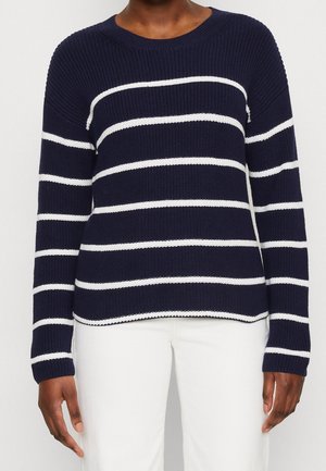 Personne portant un pull en maille bleu marine avec des rayures blanches horizontales et un pantalon blanc, debout sur un fond uni clair.