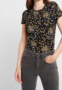 Camiseta negra con flores pequeñas en blanco y amarillo, de mangas cortas y escote redondo, combinada con unos vaqueros de talle alto grises.