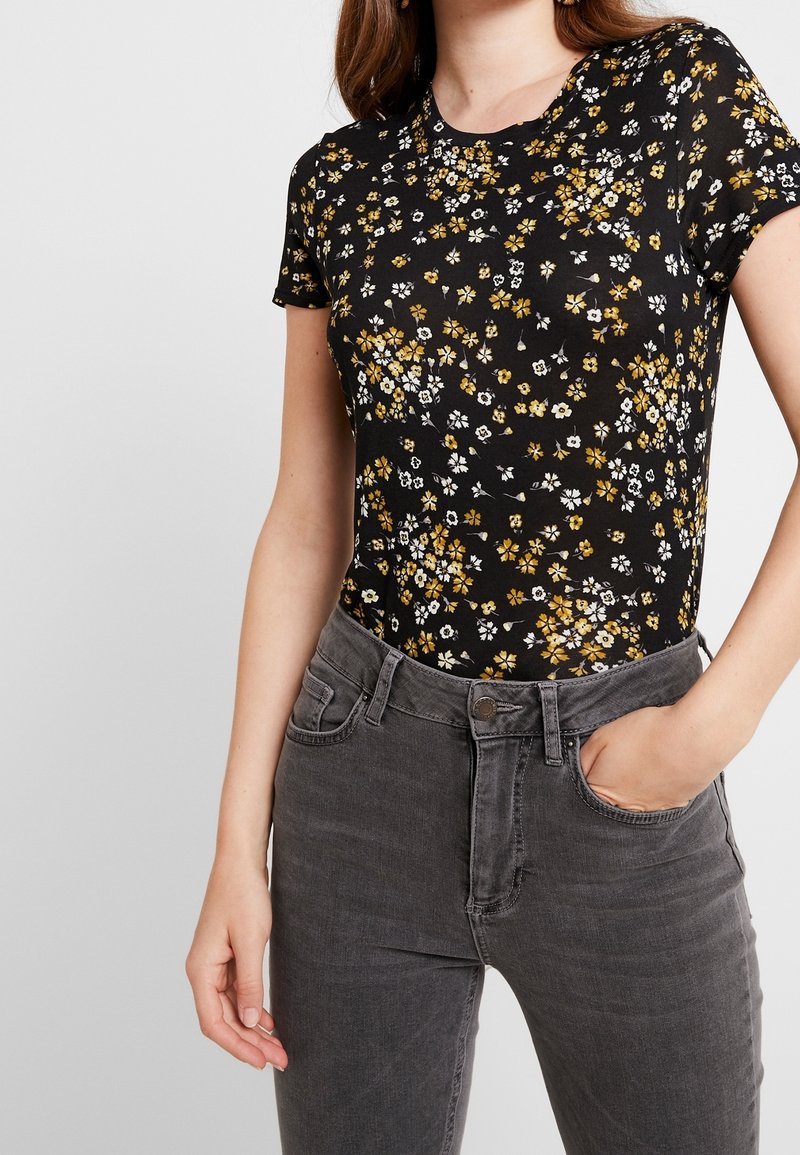 Camiseta negra con flores pequeñas en blanco y amarillo, de mangas cortas y escote redondo, combinada con unos vaqueros de talle alto grises.