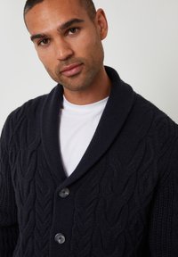 Cárdigan de punto azul marino con cuello de chal, que presenta un patrón de punto trenzado y tres botones oscuros en la parte delantera, superpuesto a una camiseta blanca.