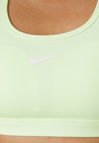 Soutien-gorge de sport vert clair en tissu extensible, présentant un col rond et un logo Nike blanc à l'avant. Texture lisse et design ajusté.