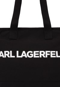 Sac cabas en tissu noir avec un logo blanc "CARL LAGERFELD" bien visible sur le devant, équipé de deux poignées robustes pour le transport.