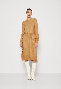 Rochie tricotată într-o nuanță caldă de portocaliu, cu mâneci lungi, talie accesorizată cu o curea și slituri laterale. Asortată cu cizme albe până la genunchi.