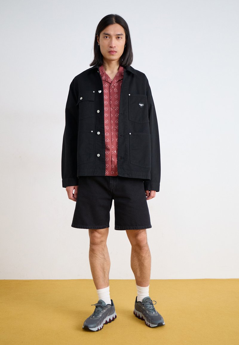 Chaqueta negra con bolsillos, camisa roja con estampado, pantalones cortos negros, zapatillas grises con detalles rojos. Tejidos texturizados, ajuste casual, diseño contemporáneo.