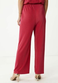 Wijde beenbroeken van felrode stof met een gestructureerd plooienpatroon, een elastische tailleband en gedragen met metallic hoge hakken.