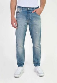 Jean en denim bleu clair à coupe slim, présentant des détails usés et un léger délavage. Associés à des baskets blanches, au design minimaliste.