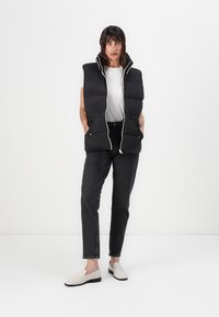 Svart puffervest med høy krage, hvit glidelås, og to frontlommer, sammen med en hvit t-skjorte og mørke jeans, iført lyse sko.