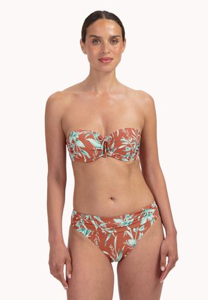 Frau trägt ein rostfarbenes, geblümtes Bandeau-Bikini-Set und steht mit entspannten Armen vor einem schlichten weißen Hintergrund.