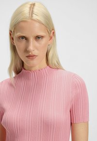 Różowa, prążkowana bluzka z krótkimi rękawami i wysokim dekoltem. Posiada delikatnie postrzępione krawędzie oraz mały detal z logo na rękawie.