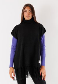 Poncho nero in lana con collo alto, caratterizzato da maniche corte e un bordo frangiato. Sotto, si intravede una maglietta viola a maniche lunghe.