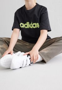 Mustapaita, jossa on lyhyet hihat ja vihreä "adidas" -logo, yhdistettynä teksturoituihin ruskeisiin housuihin ja valkoisiin tennareihin, joissa on tarranauhat.