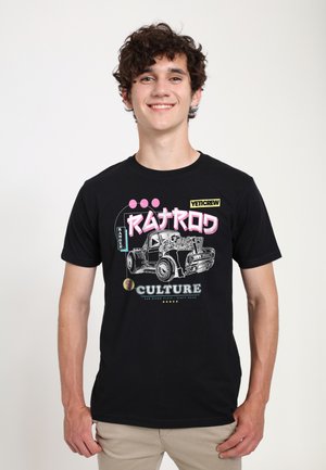 Smilende ung mand med krøllet hår iført en sort T-shirt med en vintage lastbil illustration og teksten "Ratrod Culture" og "YetiCrew".